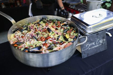 Large pan of mixed seafood and sausage paella on a catering table with a serving tray and sign reading "Briar College Catering.