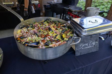 Large pan of seafood paella with mussels, sausage, and vegetables on a catering table next to a covered tray.