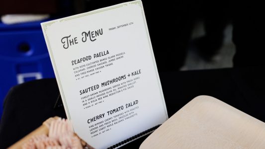 A printed menu lists seafood paella, sautéed mushrooms with kale, and cherry tomato salad on a counter.
