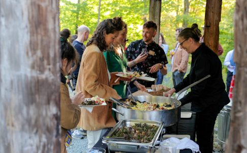 A group of people stands in line outdoors as a woman serves food from a large pan at a buffet-style table.