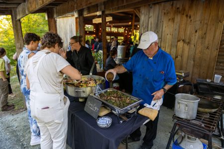 A chef serves food from chafing dishes to people at an outdoor buffet next to a wooden building.