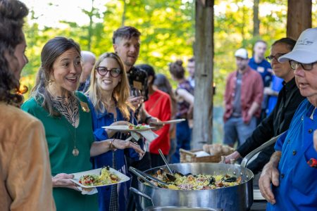 People stand in line outdoors, holding plates, as food is served from a large pot by someone in a blue chef’s coat.