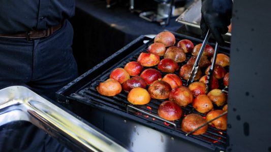 A person uses tongs to grill halved peaches on a barbecue grill.