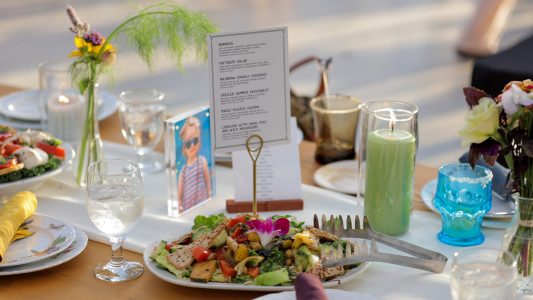 A table set with a salad platter, menu, water glasses, candles, and floral arrangements at a meal gathering.