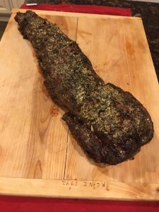 A cooked, herb-crusted beef tenderloin rests on a wooden cutting board.