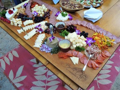 A wooden board with assorted cheeses, cured meats, fruits, nuts, and dips, arranged on a table with a red patterned rug.