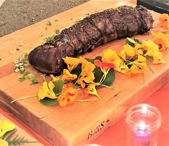 A cooked beef tenderloin on a wooden cutting board, garnished with edible flowers and chopped green onions.
