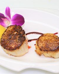 Two seared scallops on a white plate with a purple orchid, lemon wedge, and decorative sauce.