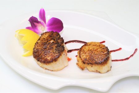 Two seared scallops on a white plate with a purple orchid, lemon wedge, and decorative sauce.