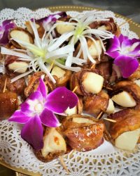 Bacon-wrapped dates with cheese on a lace doily, garnished with purple orchids and white flowers.