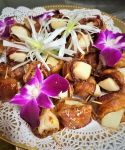 Bacon-wrapped dates with cheese on a lace doily, garnished with purple orchids and white flowers.