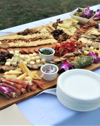 A large charcuterie board with assorted cheeses, meats, crackers, fruit, dips, and plates arranged on a white tablecloth.
