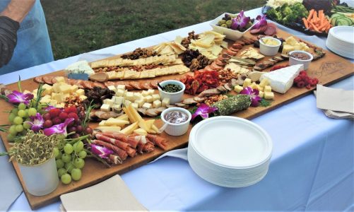 A large charcuterie board with assorted cheeses, meats, crackers, fruit, dips, and plates arranged on a white tablecloth.