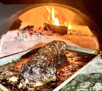 A cooked roast sits on a metal tray inside a wood-fired oven with flames in the background.