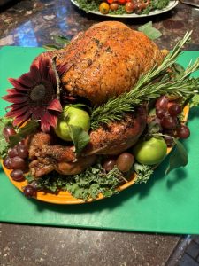 Roast turkey garnished with green apples, grapes, herbs, and a large red flower on a green cutting board.