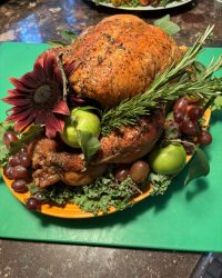 Roast turkey garnished with green apples, grapes, herbs, and a large red flower on a green cutting board.