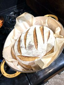 Round loaf of sourdough bread with slashes on top, baked in a parchment-lined Dutch oven on a stovetop.