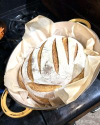 Round loaf of sourdough bread with slashes on top, baked in a parchment-lined Dutch oven on a stovetop.