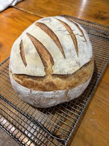 A round loaf of artisan bread with scored crust rests on a cooling rack atop a wooden surface.