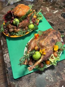 Two roasted poultry dishes garnished with fresh fruits, greens, herbs, and edible flowers on a kitchen counter.