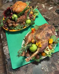 Two roasted poultry dishes garnished with fresh fruits, greens, herbs, and edible flowers on a kitchen counter.