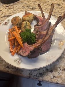 Three lamb chops with roasted potatoes, glazed carrots, and parsley garnish on a white plate on a granite countertop.