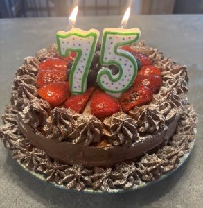 Chocolate cake with strawberries, chocolate frosting, and lit "75" candles on top, sitting on a plate.