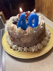 Round cake with whipped cream and berries, topped with lit blue candles shaped as the number 40 on a yellow plate.