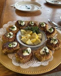 A platter of crab cakes topped with sauce and herbs, arranged around a bowl of lemon wedges on a gold plate.