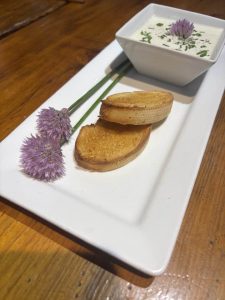 A white plate with toasted bread slices, a bowl of creamy dip, and two purple chive flowers as garnish.
