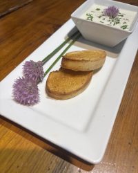 A white plate with toasted bread slices, a bowl of creamy dip, and two purple chive flowers as garnish.
