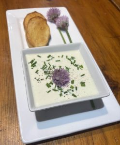 A square bowl of creamy soup garnished with chives and a purple flower, served with toasted bread slices on a white plate.