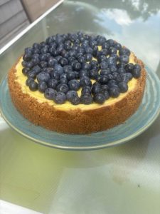 A cheesecake with a graham cracker crust topped with fresh blueberries sits on a blue plate on a glass table.