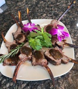 Plate of cooked lamb chops garnished with mint, rosemary, and purple flowers on a white square plate.