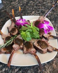 Plate of cooked lamb chops garnished with mint, rosemary, and purple flowers on a white square plate.
