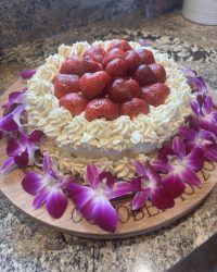 Round cake topped with whole strawberries and cream, surrounded by purple orchids on a wooden board.