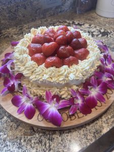 Round cake topped with whole strawberries and cream, surrounded by purple orchids on a wooden board.