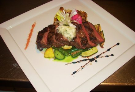 Sliced steak with herb butter on sautéed vegetables, garnished with greens and sauce on a white square plate.