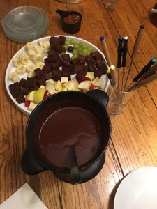 A fondue pot filled with chocolate is next to a platter of fruit and brownie bites, with fondue forks on the side.