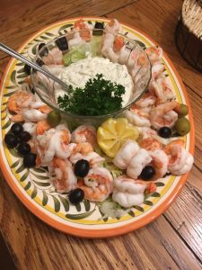 Shrimp arranged around a bowl of dip, garnished with parsley, lemon, black and green olives on a decorative platter.