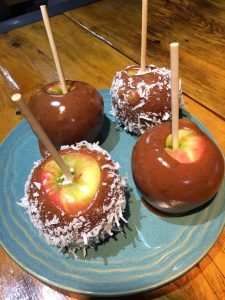 Four candy apples on a blue plate, two coated with shredded coconut, on a wooden table.