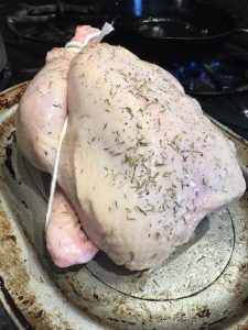 Raw whole chicken seasoned with herbs and tied with string, placed on a roasting pan before cooking.