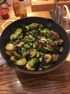 A skillet filled with roasted Brussels sprouts drizzled with balsamic glaze sits on a wooden table.