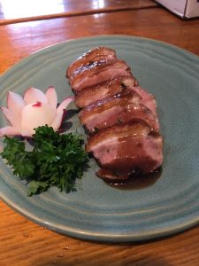 Slices of cooked duck breast with sauce, garnished with parsley and a radish carved like a flower on a blue plate.