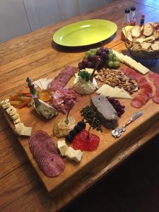 A wooden board with assorted meats, cheeses, fruits, spreads, and nuts sits on a wooden table beside a plate and crackers.