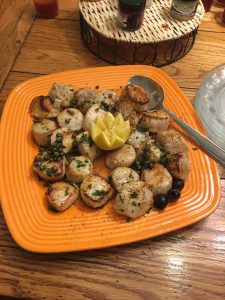 Seared scallops garnished with herbs and spices on an orange plate, with a lemon wedge and a few black olives.
