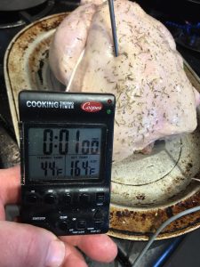 A hand holds a cooking thermometer showing 44°F next to a raw seasoned turkey with a probe inserted.