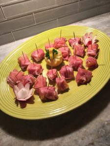 A green platter holds prosciutto-wrapped appetizers on toothpicks, with a lemon garnish and radish flowers.