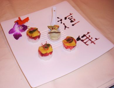 A square white plate with three sushi pieces, a dollop of wasabi, and a purple orchid, decorated with sauce writing.