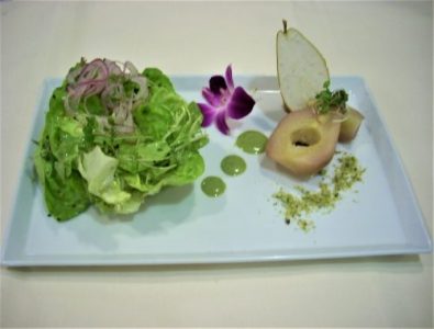A plated salad with lettuce, sliced red onions, poached pear, green sauce dots, crushed nuts, and a purple flower garnish.